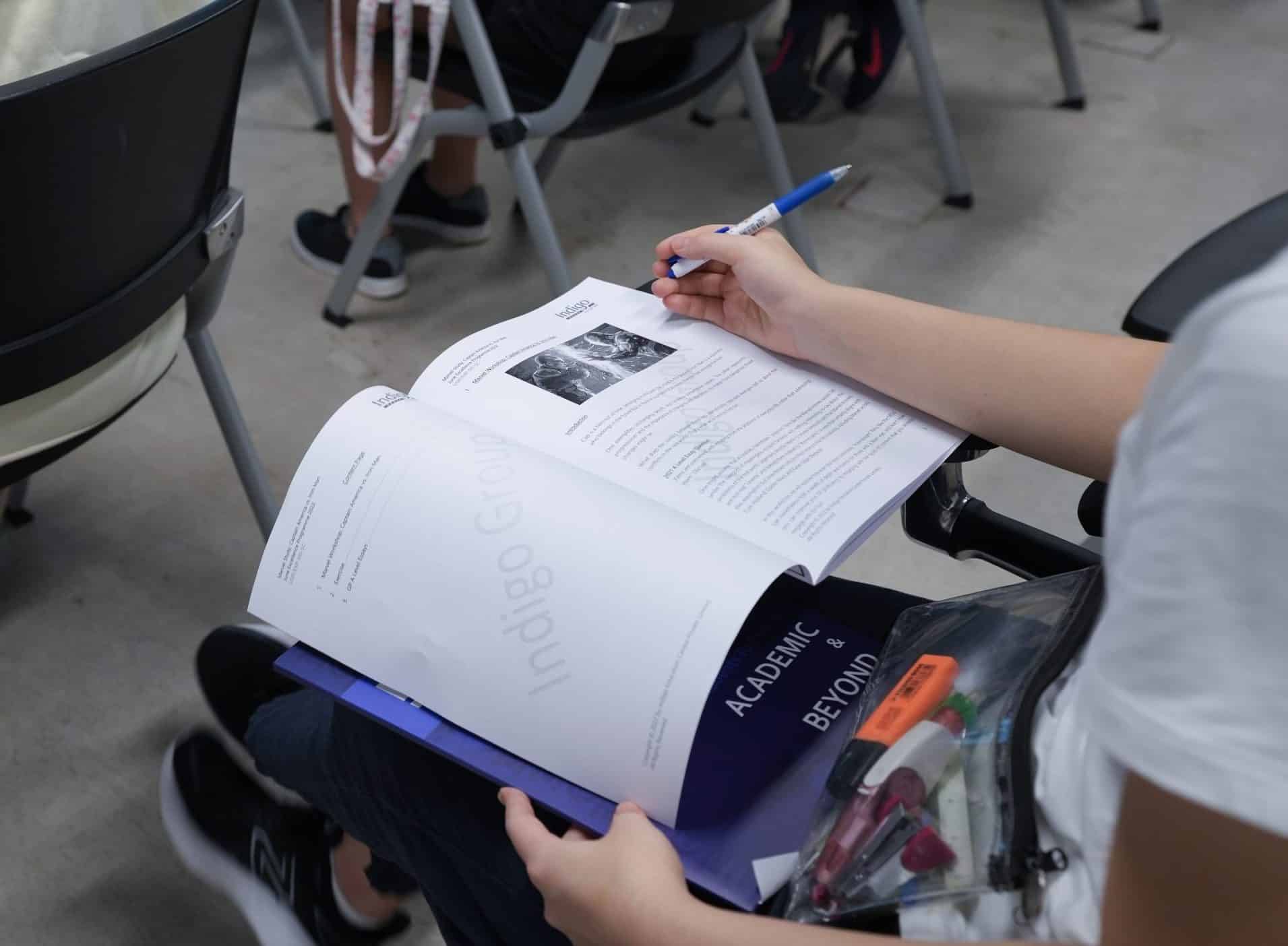 A student holding a pen with a workbook on the table before them.