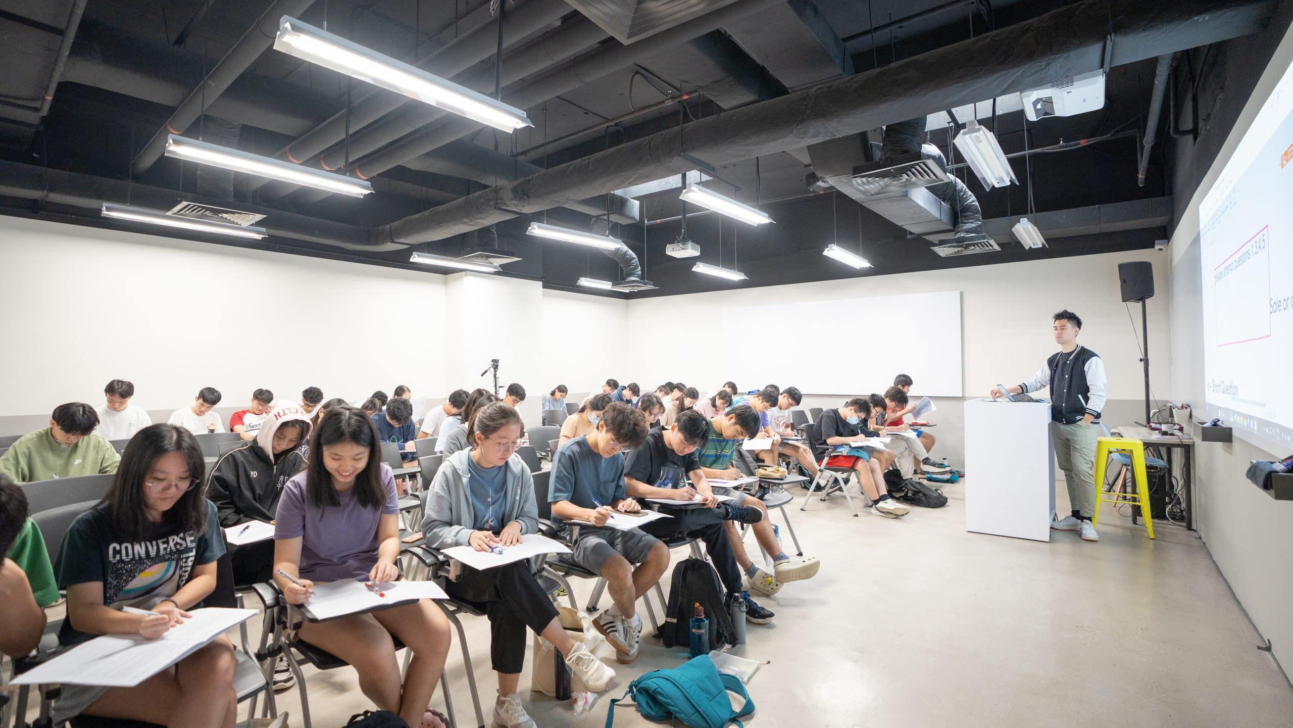 A tuition lesson takes place in a classroom with students seated in lecture seats as a tutor conducts the class at the front.